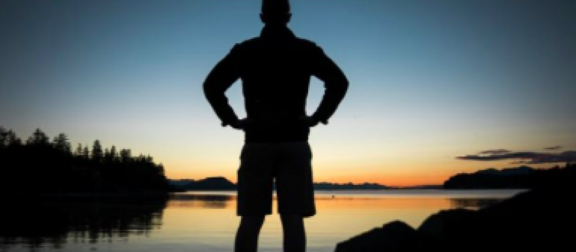 [Image description: Silhouette of a figure in shorts standing in front of a body of water, their hands on their hips. It looks to be sunrise or sunset. Image by Steve Halama on Unsplash.]